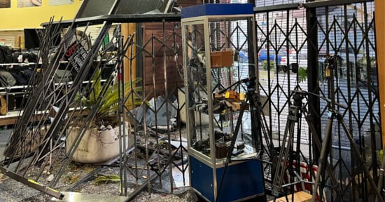 A store interior with metal security grilles bent and glass cases damaged, displaying signs of a break-in. Shattered glass covers the floor alongside fallen plants and camera equipment.