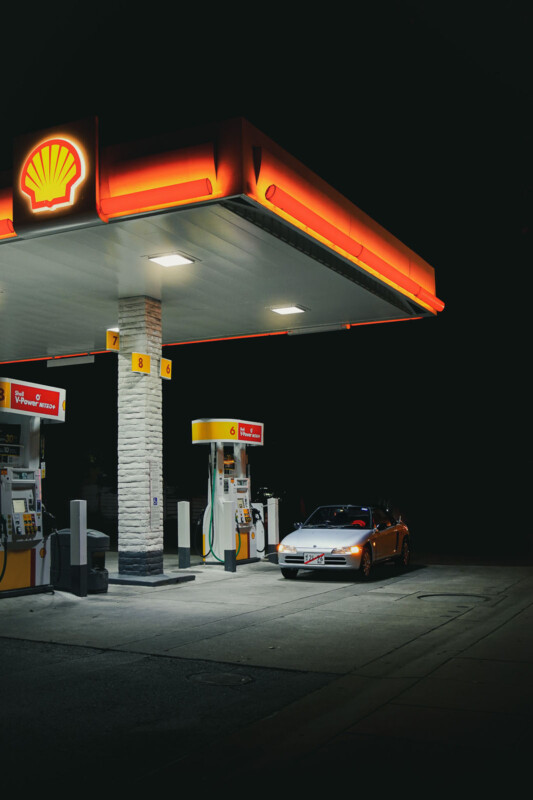 A car is parked at a Shell gas station at night. The illuminated canopy displays the Shell logo, casting a warm glow on the vehicle and the surrounding area. The scene is mostly dark, highlighting the gas station lights.