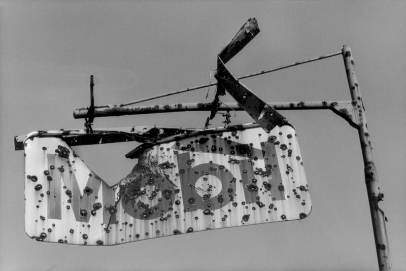 A heavily weathered and bullet-riddled sign with partial letters "MO" is attached to a rusted metal pole against a plain sky. The sign appears old and worn, suggesting abandonment.