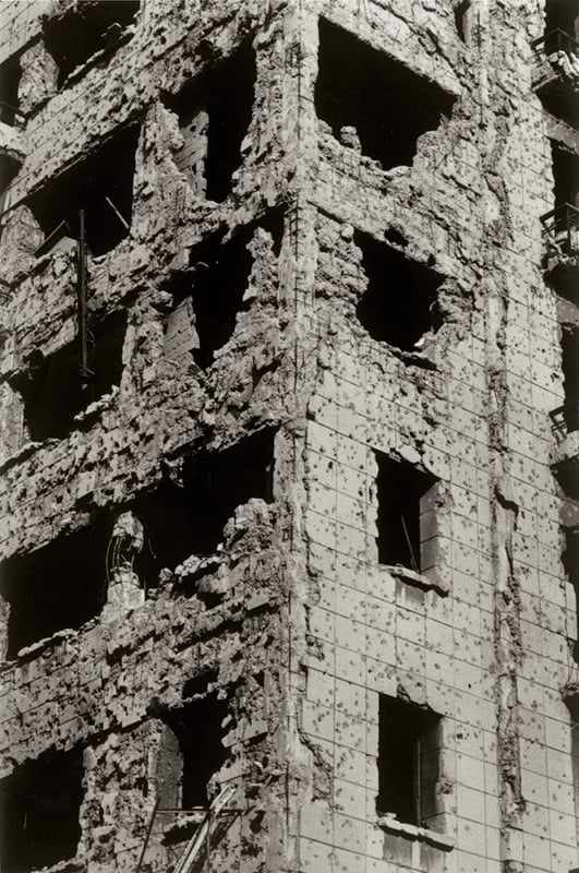 A tall, heavily damaged concrete building with numerous holes and crumbling sections. The structure shows signs of severe destruction, likely from conflict or disaster, exposing parts of the interior. The overall atmosphere is one of devastation.