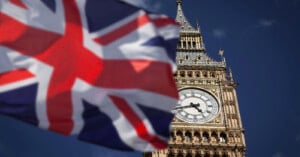 The image shows a close-up of Big Ben's clock tower against a clear blue sky. A waving, partially blurred Union Jack flag is in the foreground on the left, adding a dynamic element to the scene.