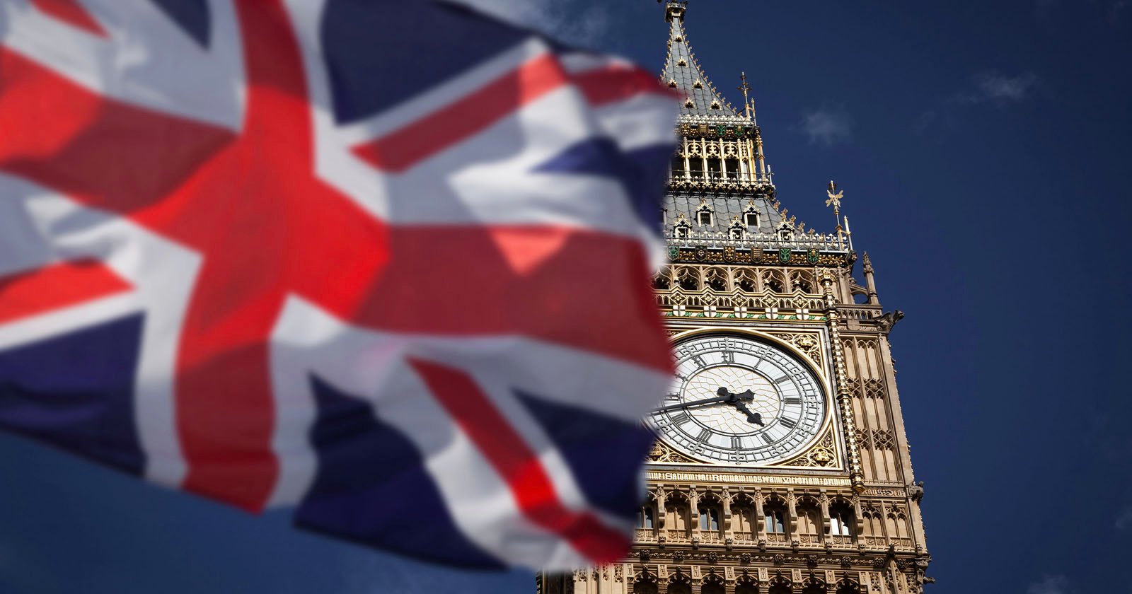 The image shows a close-up of Big Ben's clock tower against a clear blue sky. A waving, partially blurred Union Jack flag is in the foreground on the left, adding a dynamic element to the scene.