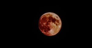 A full moon with a reddish hue is set against a dark sky. The surface of the moon is detailed, showing lunar craters and shadow contrasts. The reddish tint suggests a possible lunar eclipse or atmospheric effect.