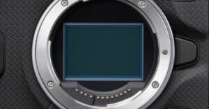 Close-up of a digital camera's sensor, surrounded by a metal mount. The camera's body has a textured black surface. The sensor is rectangular, with electronic contacts visible along the mount's inner edge.