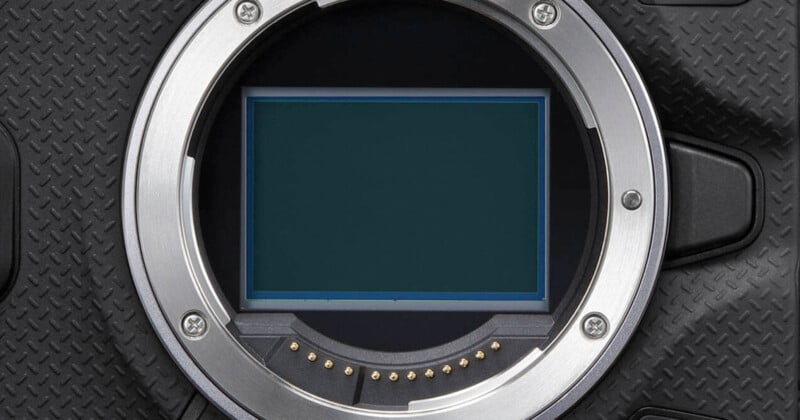 Close-up of a digital camera's sensor, surrounded by a metal mount. The camera's body has a textured black surface. The sensor is rectangular, with electronic contacts visible along the mount's inner edge.