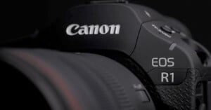 Close-up of a Canon EOS R1 camera, showcasing the brand logo and model name on a textured black body, with part of the attached lens visible on the left.