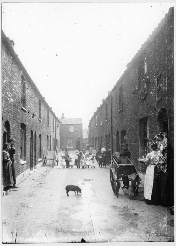 Black and white photo of a narrow street with terraced houses. Children play in the center, and adults stand by doorways. A man pushes a cart past a pig in the foreground. Laundry hangs across the alley under an overcast sky.