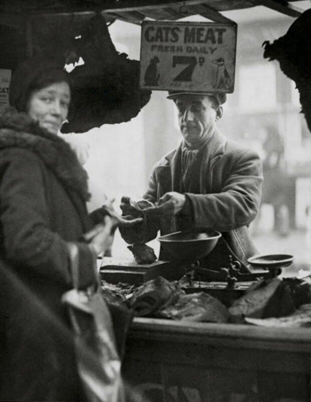 A black-and-white photo shows a man behind a market stall labeled "Cats Meat Fresh Daily," handing meat to a woman in a coat and hat. Various meats are displayed on the counter in front of him.