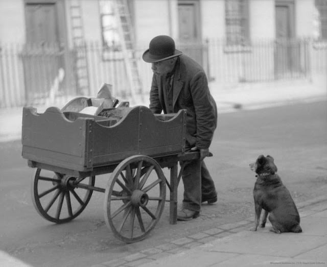 A man in a bowler hat leans over a wooden cart filled with objects on a cobblestone street. A small dog sits nearby, looking attentively at the man. A row of buildings and a ladder are in the background. The scene is in black and white.