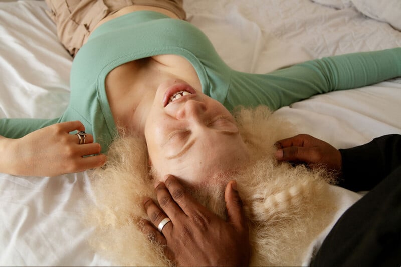 A person with fluffy blonde hair lies on their back, eyes closed, smiling. They wear a light green top. Another person's hands gently cradle their head, creating a calm and soothing atmosphere.