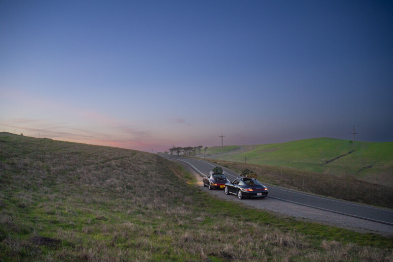 A car is driving down a rural road at sunset, towing a trailer with a large Christmas tree. The landscape is expansive with rolling green hills and a clear evening sky transitioning from orange to deep blue.