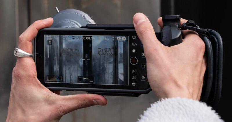 A person holding a camera with a large lens and viewing screen. The screen displays a close-up image of graffiti on a wall. The person's other hand adjusts the lens focus, and a ring is visible on one finger. They're wearing a white, fuzzy sleeve.