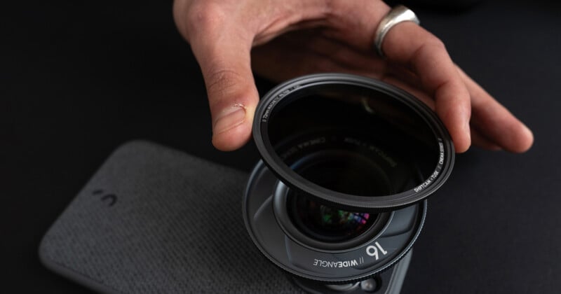 A hand with a silver ring is holding a camera lens filter above a smartphone with an attached wide-angle lens. The filter is circular, and the smartphone has a textured back with a small "u" logo. The background is dark.