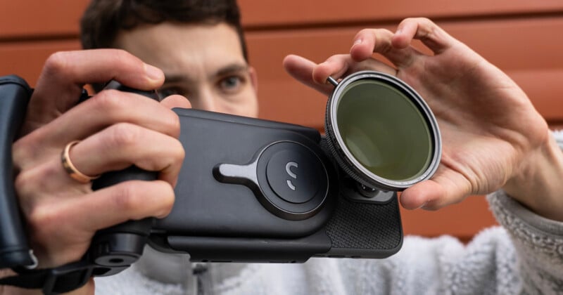 A person adjusts a circular camera lens filter attached to a black camera rig, standing in front of a red, textured wall. They have short hair and are wearing a light gray jacket. Their fingers are spread to hold the filter securely.