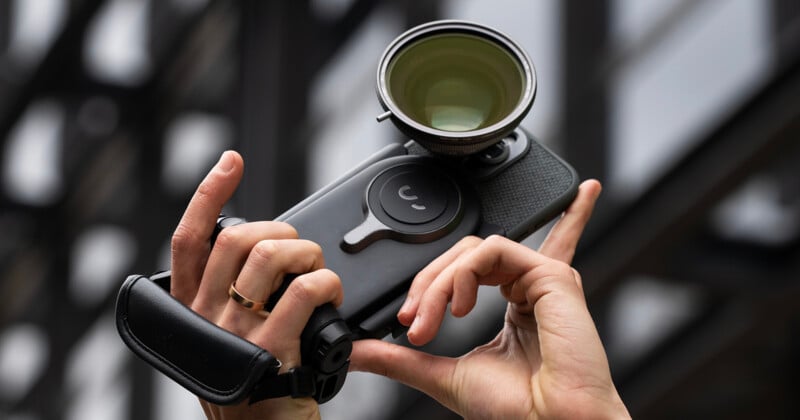 Close-up of hands holding a professional camera with an attached large lens and a hand strap. The background features blurred structural elements, suggesting an outdoor setting.