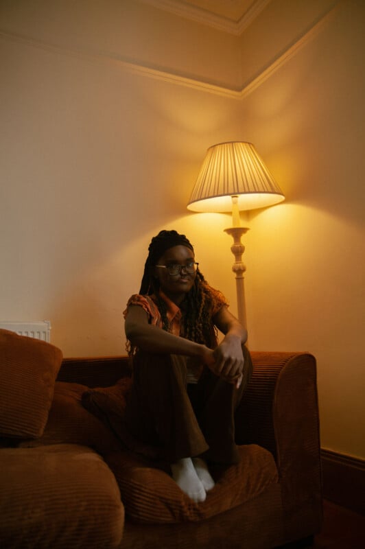 A person with long hair and glasses sits on a brown couch, illuminated by a warm floor lamp. They are wearing a brown outfit and white socks, with their arms resting on their knees. The setting appears cozy and softly lit.