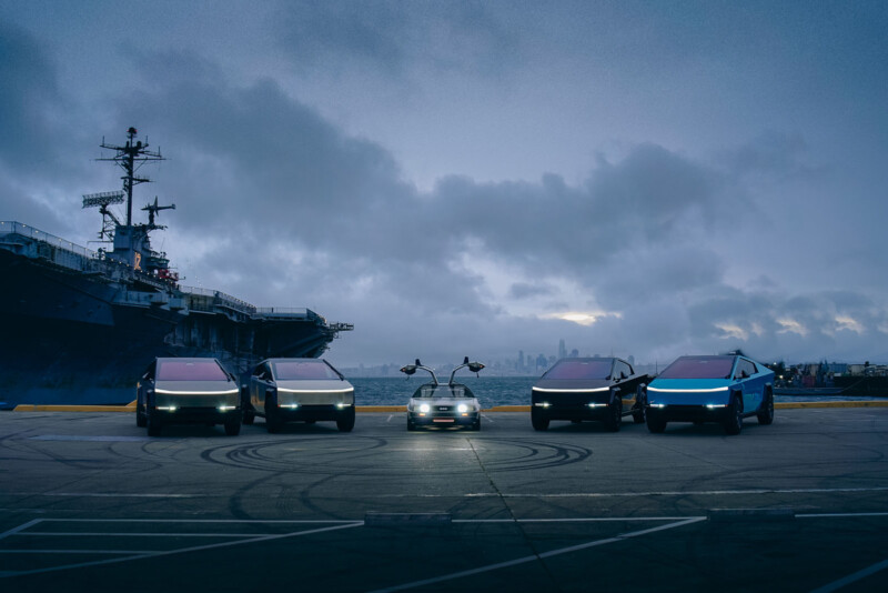 A group of futuristic cars are parked in a row near a waterfront with a large ship on the left. The vehicles have illuminated front lights and sleek exteriors. The skyline is visible in the distance under a cloudy sky.