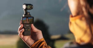 A person outdoors holds a handheld camera device with a small screen showing a video of a woman. The person wears an orange jacket. The background is blurred, featuring trees and hills.