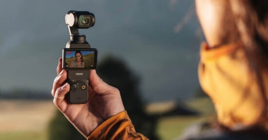 A person outdoors holds a handheld camera device with a small screen showing a video of a woman. The person wears an orange jacket. The background is blurred, featuring trees and hills.