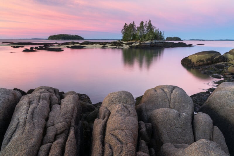 A serene coastal scene at sunset with smooth, pink-tinted water. Rugged rocks lead to a small island with dense trees. The sky is painted in soft pastel hues of pink and purple, creating a tranquil atmosphere.