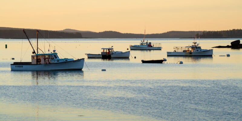 A calm seascape at sunrise with several small fishing boats anchored on the still water. The horizon is softly lit with golden light, and distant land silhouettes are visible. The serene scene reflects warm hues on the water's surface.