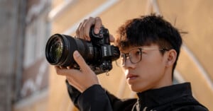 A person wearing glasses and a black jacket is holding a Nikon camera, focusing on capturing a photo. The background features a yellow wall with architectural details.