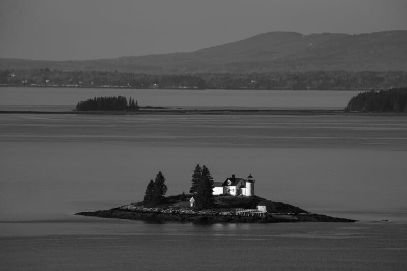 A small island with a lighthouse and two buildings, surrounded by calm water. A few trees stand on the island. In the background, distant hills and forests are visible under a clear sky. The image is in black and white.