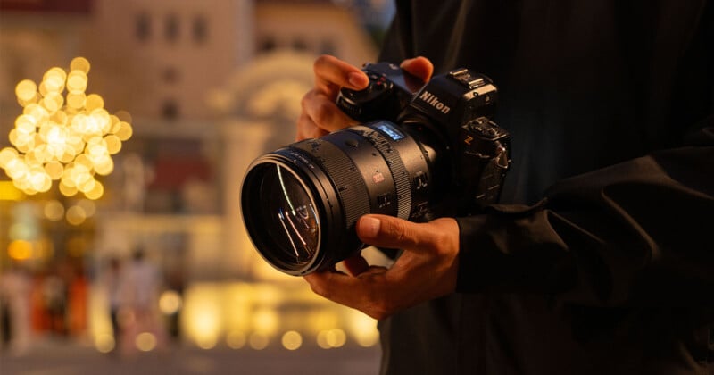 A person holding a Nikon camera with a large lens, standing outdoors in a softly lit urban environment. Blurred warm lights and buildings create a bokeh effect in the background.