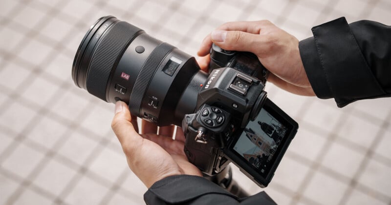 A person holding a DSLR camera with a large lens attached, pointing at the ground. The camera's LCD screen is flipped out, displaying a photo. The person is wearing a black jacket, and the background is a tiled surface.