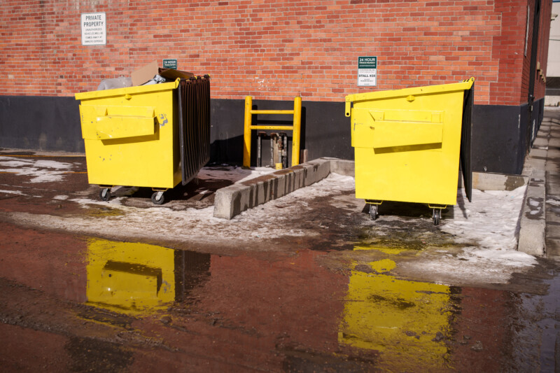 Two yellow dumpsters stand on a slightly wet, snowy area against a red brick wall. Reflections of the dumpsters are visible in the water on the ground. Signs labeled "Private Property" and caution signs are on the wall.