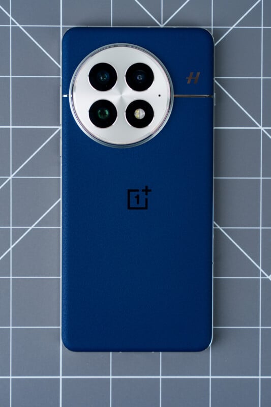 A blue smartphone with a large circular camera module featuring three lenses is placed on a grid-patterned surface. The phone has a logo in the center and minimalist design elements.