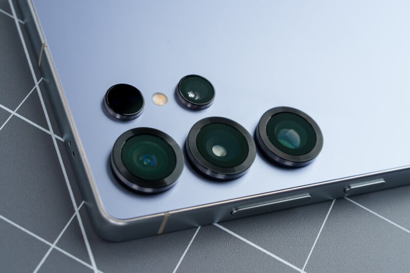 A close-up of a silver smartphone's back, showing four camera lenses and a flash. The phone is placed on a surface with a geometric pattern.