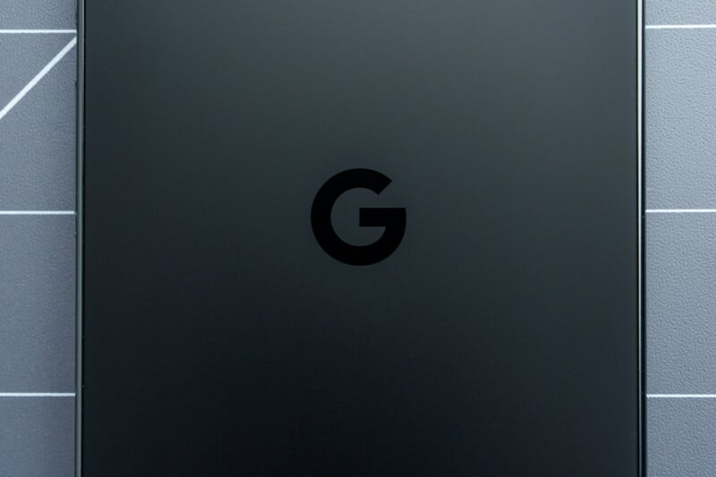 Close-up of a sleek, matte black smartphone featuring the Google "G" logo on the back. The background shows a textured surface with white grid lines.