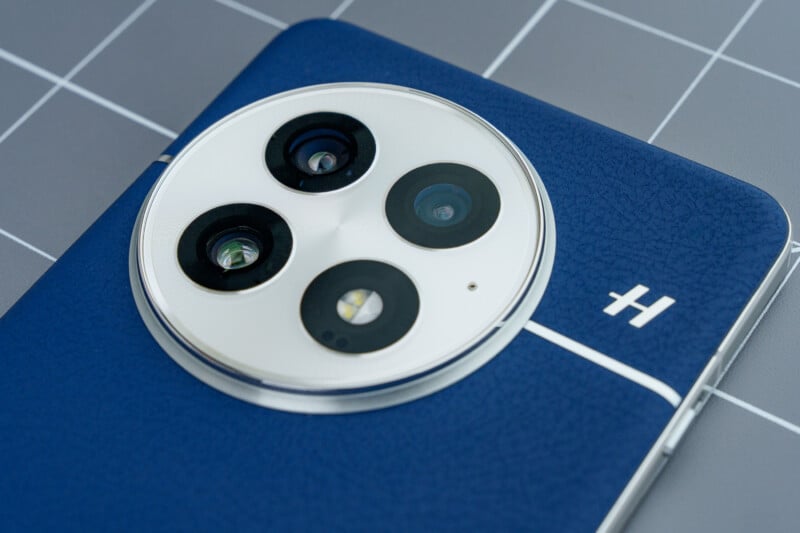 Close-up of a blue smartphone back with a large, round triple camera module and small flash. The phone is on a gray, grid-patterned surface. The back material appears textured.