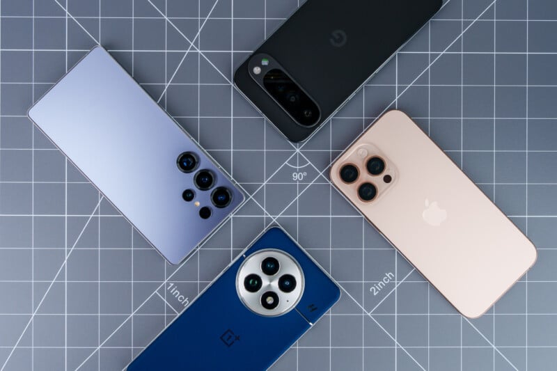 Four smartphones are arranged in a circle on a gray grid mat. The phones feature different colors and camera designs, including a light blue, a peach-colored, a black, and a dark blue device, each showcasing its unique camera layout.