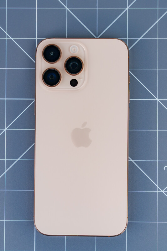 A gold smartphone with three camera lenses on the back, placed on a grid-patterned surface. The Apple logo is visible on the back of the phone.