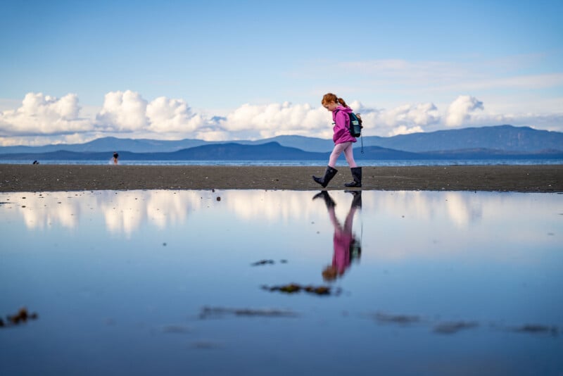 A girl in a pink jacket and boots walks beside a reflective tidepool on a sandy beach. The sky is blue with scattered clouds, and mountains are visible in the background.