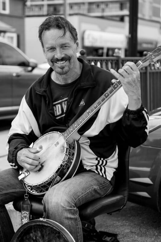 A man sits on a chair outdoors, playing a banjo and smiling. He wears a zip-up jacket and jeans. Cars and a building are visible in the background. The image is in black and white.