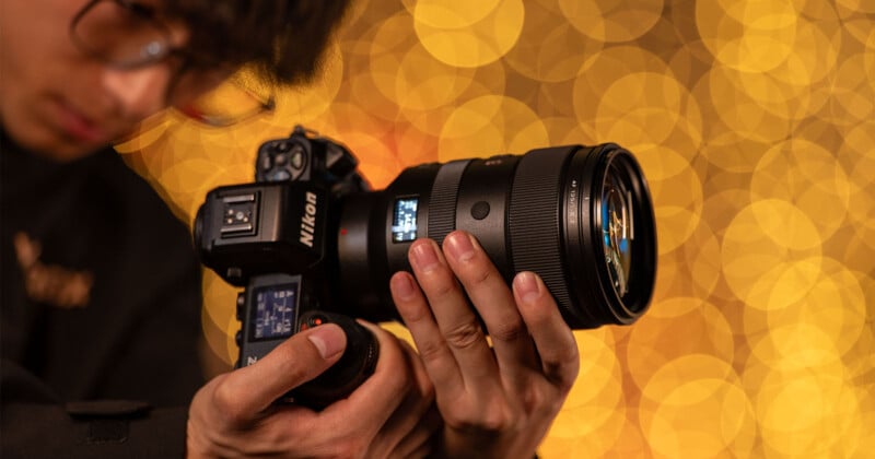 A person adjusts a Nikon camera, focusing the lens. The background features a warm bokeh effect with circular yellow lights, creating a cozy ambiance. The person wears glasses and is concentrated on the camera settings.