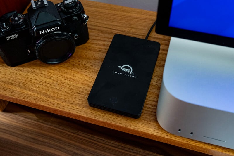 A wooden desk with a Nikon camera, a black OWC Envoy Ultra external hard drive, and the corner of a computer monitor and Mac Studio visible. Cables are attached to the devices.