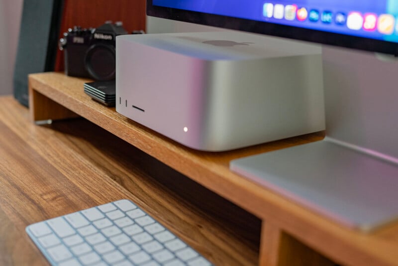 A desktop setup featuring a Mac Studio on a wooden desk, accompanied by a sleek keyboard and a small digital camera in the background. The computer screen displays vibrant icons, suggesting a macOS interface.