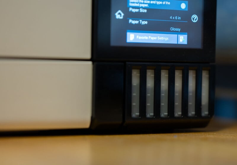 Close-up of a digital printer screen displaying settings for paper size and type, with options for 4 x 6 inches and glossy paper. The printer's exterior is partially visible, set on a wooden surface.