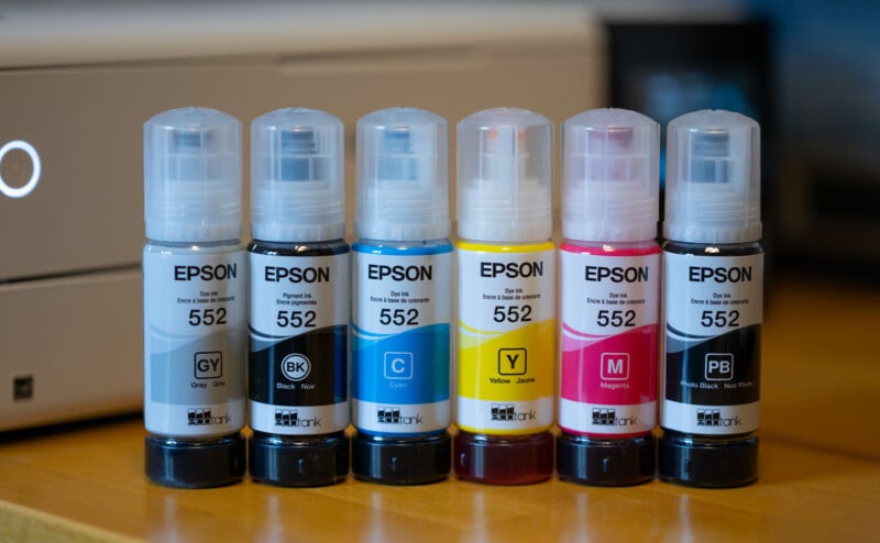 Six Epson 552 ink bottles are lined up. They include gray, black, cyan, yellow, magenta, and photo black inks. A blurred printer is visible in the background on a wooden surface.