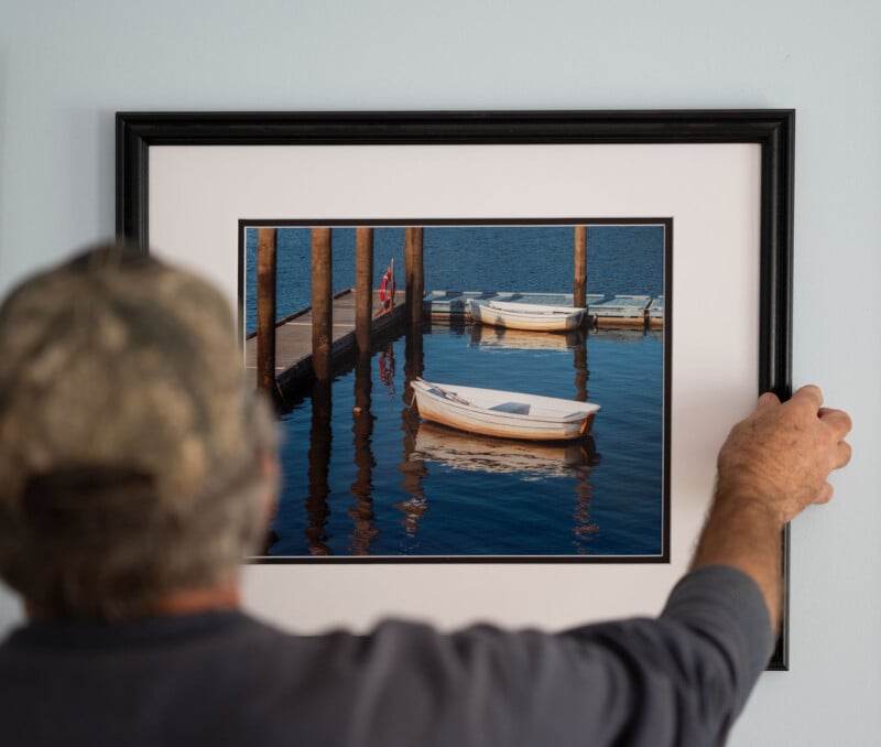 A person hangs a framed photograph on a wall. The photo depicts a wooden boat floating on still water at a dock, with several other boats in the background. The person's left hand is partially visible, holding the frame.