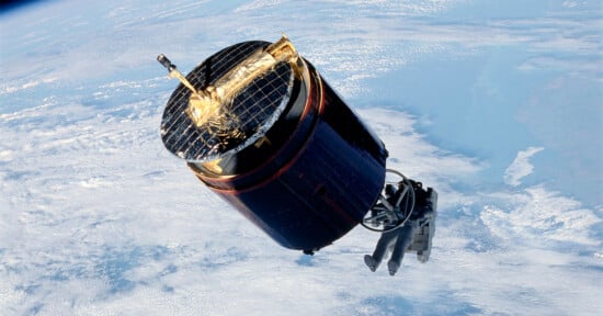 A satellite orbits Earth with solar panels glinting in the sunlight. The blue and white surface of the planet is visible in the background, highlighting the satellite's intricate design and components against the vastness of space.