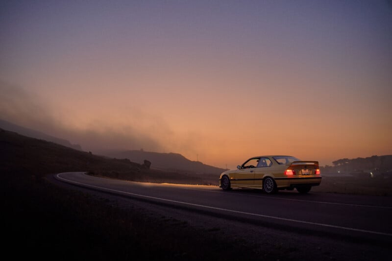 A yellow car drives along a curving road at dusk, with a misty landscape and mountains in the background under an orange and purple sky.