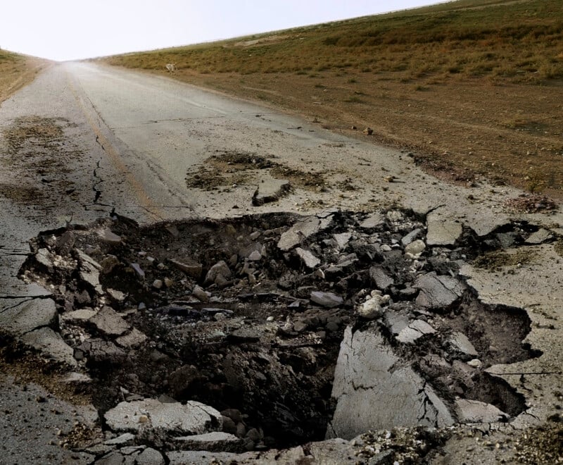 A large, deep pothole on a cracked, deserted road stretches to the horizon. The surrounding terrain is dry and barren, with patches of sparse grass along the roadside. The sky is overcast, giving a bleak atmosphere.