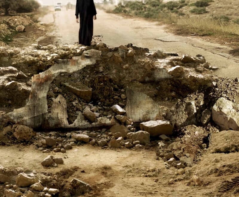 A person in dark clothing walks along a cracked, uneven road filled with rubble and debris. The scene is desolate, with dry soil and sparse vegetation surrounding the road under a cloudy sky.