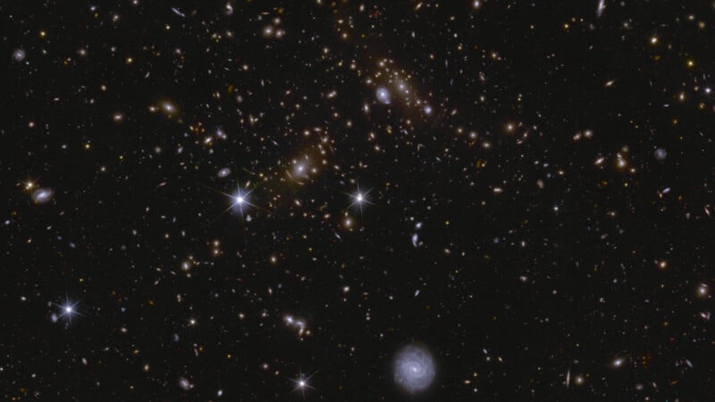 A deep space image showing numerous galaxies and stars scattered across the blackness of space. A bright spiral galaxy is visible in the lower right, with twinkling stars and dense cosmic clusters throughout the scene.