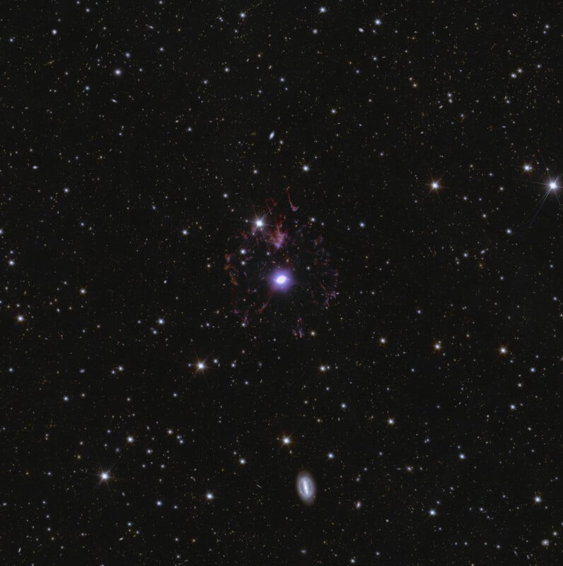 A deep space image showing a luminous galaxy at the center surrounded by faint, stringy red filaments. The background is filled with numerous stars and distant galaxies on a dark sky.